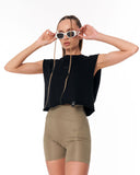 A woman modeling a black sleeveless crop top and olive green fitted biker shorts against a plain white background, holding a pair of white-framed sunglasses with a gold chain.