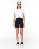 A model stands against a plain white studio background wearing a white sleeveless cropped top, black fitted biker shorts, white sneakers, and small gold hoop earrings.