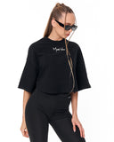 A woman modeling a black cropped t-shirt with a white script logo on the chest, paired with black leggings, against a plain white background.