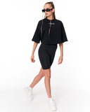 A model wearing a black short-sleeve crop top with a gold zipper and a small white logo, paired with black bike shorts and white sneakers, on a white studio background.