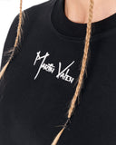 Close-up of a black crewneck sweatshirt featuring a white embroidered 'Martin Valen' logo on the chest, worn by a model with blonde braids.