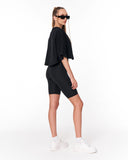 A woman modeling a black cropped t-shirt and black biker shorts, styled with white sneakers, white socks, and black sunglasses against a plain white studio background.