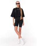 A woman modeling a black two-piece crop top and biker shorts set with white sneakers and sunglasses against a plain white background.
