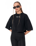 A model wearing a black cropped t-shirt with a white script logo on the chest, paired with black leggings and black sunglasses, set against a plain white background.