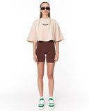 A model stands against a white studio background wearing a light beige cropped t-shirt with black text, dark brown fitted biker shorts, green and white sneakers, and black sunglasses.