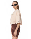 A woman modeling an oversized light beige cropped T-shirt with a small black graphic on the chest and sleeve, paired with brown fitted shorts against a plain white background.