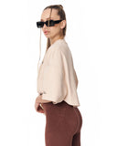 A woman in profile wearing a light beige cropped tunic top with wide sleeves, brown leggings, and black sunglasses against a plain white background.