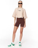 A model wearing a beige cropped t-shirt with a black graphic, dark brown bike shorts, white socks, and green and white sneakers, standing against a plain white background.