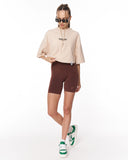 A model wearing a beige cropped T-shirt with 'Martin Valen' printed on the chest, brown fitted bike shorts, and green and white sneakers, posing against a solid white background.