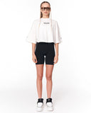 A model stands against a white studio background wearing a white cropped t-shirt with 'Martin Valen' printed on the chest, black bike shorts, and white sneakers.