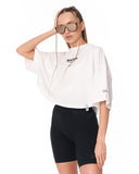 A woman modeling a white oversized graphic T-shirt with black bike shorts, sunglasses, and a gold chain necklace against a plain white background.