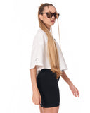 A woman wearing a white short-sleeve cropped t-shirt and black biker shorts, styled with brown cat-eye sunglasses and a long chain accessory, photographed against a solid white background.