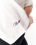 Close-up of a person wearing a white short-sleeve top with a black cursive signature-style logo on the sleeve, paired with black bottoms, against a white background.