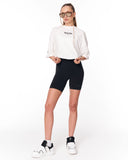 A model wearing a white cropped sweatshirt with 'Martin Valen' printed on the chest, black bike shorts, and white sneakers with black accents, standing against a plain white background.