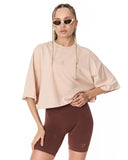 A woman modeling an oversized short-sleeve crop top in light beige with a small tonal chest logo, paired with dark brown fitted biker shorts, against a plain white background.