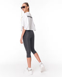 A woman modeling a white cropped t-shirt with black text on the back and dark gray capri leggings, paired with white sneakers, photographed from the back against a white studio background.