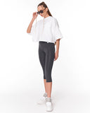 A woman modeling a white short-sleeve cropped top and dark gray capri leggings with white sneakers, standing against a plain white studio background.