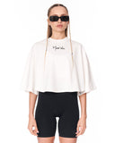 A model wearing a white cropped t-shirt with a black script logo on the chest, paired with black bike shorts and black sunglasses, against a plain white background.