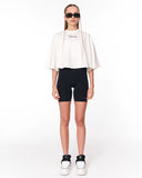 A model stands against a plain white studio background wearing a white cropped tunic top with a small black script logo on the chest, black fitted shorts, black-and-white sneakers, and black sunglasses.