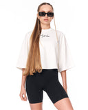 A model wearing a white cropped T-shirt with a black script logo on the chest, paired with black bike shorts and black sunglasses, against a plain white background.