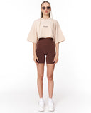 A model stands against a white studio background wearing a light beige cropped t-shirt with a small black logo on the chest, dark brown high-waisted bike shorts, white sneakers, and sunglasses.