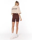 A woman is shown from behind wearing a beige cropped t-shirt with 'MARTIN VALEN' printed on the back and dark brown biker shorts, paired with white sneakers, against a plain white background.
