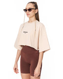 A woman modeling a cropped beige t-shirt with 'MARTIN VALEN' printed on the chest, paired with brown bike shorts, against a plain white background.