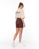 A woman is shown from the back wearing a cream cropped t-shirt with 'MARTIN VALEN' printed on the back and dark brown high-waisted biker shorts, paired with white sneakers, against a plain white background.