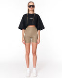 A model stands against a white studio background wearing a black cropped t-shirt with a small white logo, beige high-waisted bike shorts, white sneakers, and black sunglasses.