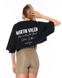 A woman shown from the back wearing a black oversized cropped T-shirt with white text reading 'MARTIN VALEN' and smaller script text below, paired with beige fitted shorts, against a plain white background.