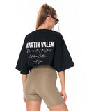 A woman shown from the back wearing a black oversized crop top with white 'Martin Valen' text on the back and beige fitted biker shorts against a plain white background.