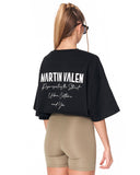 A woman shown from the back wearing a black oversized Martin Valen graphic t-shirt and beige fitted shorts against a plain white background.