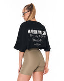 A woman shown from the back wearing a black oversized crop top with white text reading 'MARTIN VALEN' and beige fitted shorts against a plain white background.