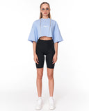 A woman modeling a light blue cropped t-shirt with a white graphic print, paired with black biker shorts and white sneakers, standing against a plain white studio background.