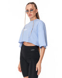 A woman modeling a light blue cropped t-shirt with a small white graphic on the chest, paired with black high-waisted biker shorts, against a plain white background.