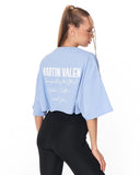 A woman shown from the back wearing a light blue cropped t-shirt with white text that reads 'MARTIN VALEN' and 'Reimagining the Streetwear Culture, and You' paired with black leggings, against a plain white background.