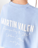 A close-up of a light blue crewneck sweatshirt featuring large white 'MARTIN VALEN' text and white script text on the front, worn by a model.