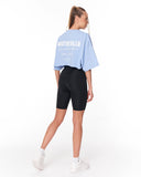 A woman shown from the back wearing a light blue cropped T-shirt with white text reading 'MARTIN VALEN' and black fitted biker shorts, paired with white sneakers, on a clean white studio background.