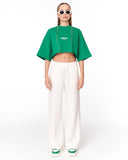 A model stands against a white studio background wearing a bright green cropped short-sleeve top with white text on the chest, paired with wide-leg white pants and green-and-white sneakers.
