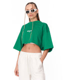 A model wearing a bright green cropped t-shirt with white text on the chest, paired with white sweatpants, against a plain white background.