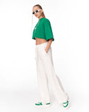 A model wearing a bright green cropped t-shirt, white wide-leg trousers, and green and white sneakers, posed against a solid white studio background.