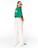 A woman modeling a green cropped T-shirt with white graphic text on the back, paired with off-white wide-leg trousers and green sneakers, photographed against a plain white studio background.