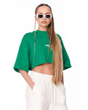 A woman modeling a cropped green t-shirt with a small white graphic on the chest, paired with white drawstring pants and white-framed sunglasses, against a plain white background.