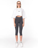 A model stands against a white studio background wearing a white cropped T-shirt with a small black chest logo, dark gray capri leggings, and white sneakers.