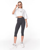 A model wearing a white cropped t-shirt with a black logo, dark gray high-waisted capri leggings, white sneakers, and sunglasses, standing against a plain white background.